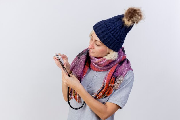 Feliz joven rubia enferma mujer eslava con sombrero de invierno y bufanda sostiene y mira estetoscopio aislado en la pared blanca con espacio de copia