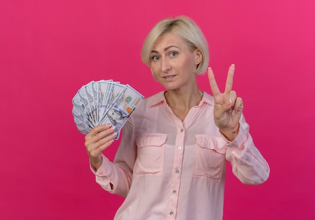 Feliz joven rubia eslava sosteniendo dinero y haciendo el signo de la paz aislado sobre fondo rosa.