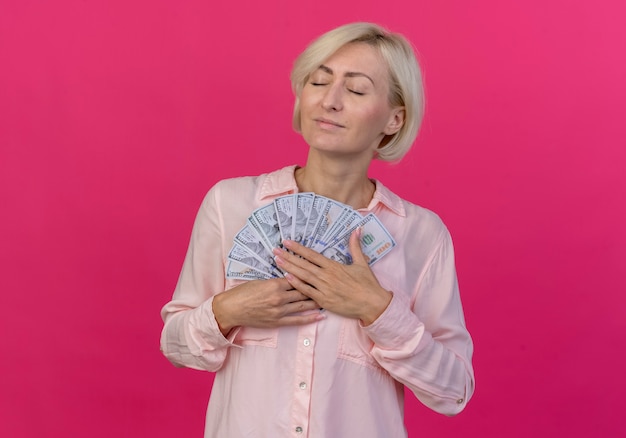 Feliz joven rubia eslava sosteniendo dinero con los ojos cerrados aislado sobre fondo rosa con espacio de copia