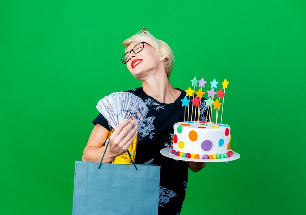 Feliz joven rubia fiestera con gafas y gorro de cumpleaños con pastel de cumpleaños con caja de regalo de dinero de estrellas y bolsa de papel sonriendo con los ojos cerrados aislado sobre fondo verde con espacio de copia