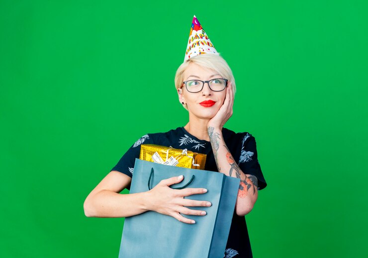 Foto gratuito feliz joven rubia fiestera con gafas y gorro de cumpleaños sosteniendo una bolsa de papel con cajas de regalo manteniendo la mano en la cara mirando al lado soñando aislado sobre fondo verde con espacio de copia