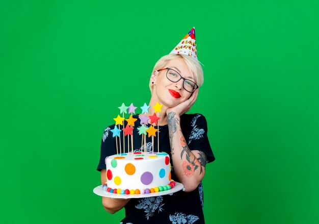 Feliz joven rubia fiestera con gafas y gorro de cumpleaños sosteniendo pastel de cumpleaños con estrellas manteniendo la mano en la cara mirando a cámara aislada sobre fondo verde con espacio de copia