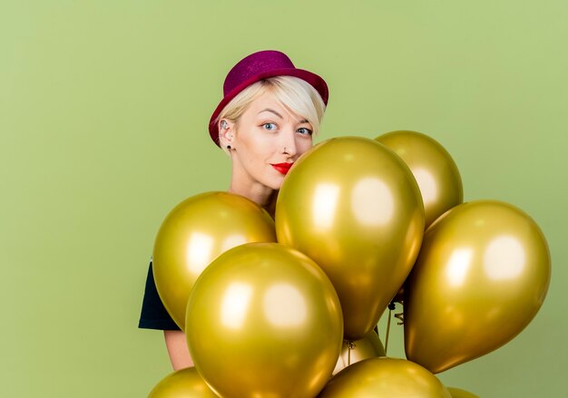 Feliz joven rubia fiestera con sombrero de fiesta de pie detrás de globos mirando a cámara aislada sobre fondo verde oliva con espacio de copia