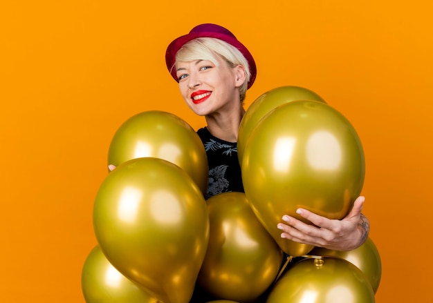 Foto gratuita feliz joven rubia fiestera con sombrero de fiesta de pie detrás de globos tocándolos mirando a cámara y sonriendo aislado sobre fondo naranja