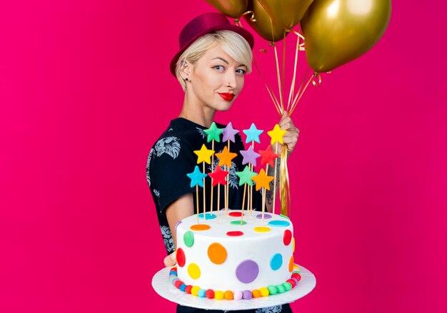 Feliz joven rubia fiestera con sombrero de fiesta de pie en la vista de perfil sosteniendo globos y estirando el pastel de cumpleaños con estrellas hacia la cámara aislada sobre fondo carmesí con espacio de copia