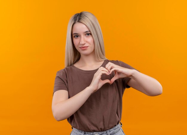 Feliz joven rubia haciendo signo de corazón mirando en espacio naranja aislado con espacio de copia