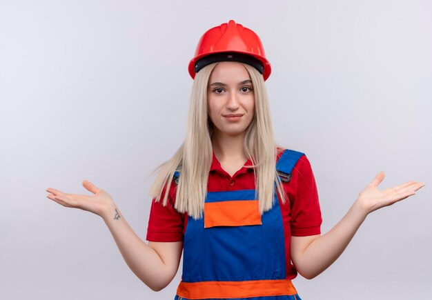 Feliz joven rubia ingeniero constructor chica en uniforme mostrando la mano vacía en un espacio en blanco aislado