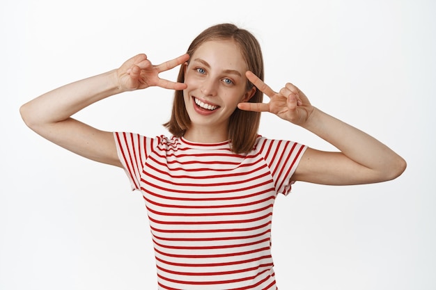 Feliz joven rubia mostrando signos de paz en v, dedos de discoteca cerca de los ojos, sonriendo con una sonrisa blanca perfecta y mirando al frente, de pie contra la pared blanca