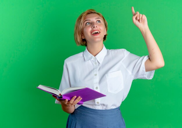 Feliz joven rubia rusa sostiene el libro mirando y apuntando hacia arriba aislado sobre fondo verde con espacio de copia