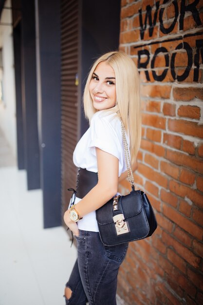 Feliz joven rubia sonriendo con la espalda y vistiendo ropa casual mirando contra la pared de ladrillo de la oficina