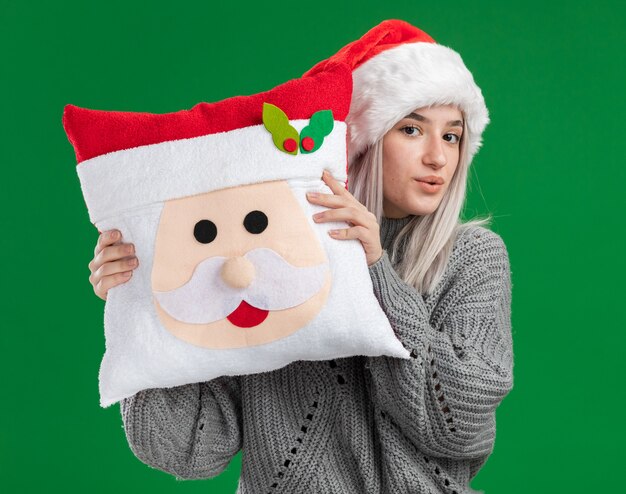 Feliz joven rubia en suéter de invierno y gorro de Papá Noel con almohada de Navidad mirando a la cámara con una sonrisa en la cara de pie sobre fondo verde