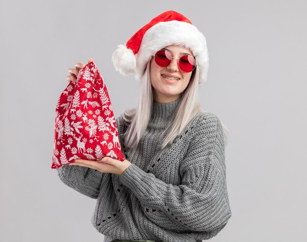 Feliz joven rubia en suéter de invierno y gorro de Papá Noel con bolsa roja de santa con regalos de Navidad sonriendo alegremente de pie sobre la pared blanca