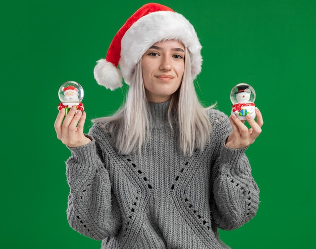 Feliz joven rubia en suéter de invierno y gorro de Papá Noel con globos de nieve de juguete de Navidad mirando a la cámara sonriendo alegremente de pie sobre fondo verde