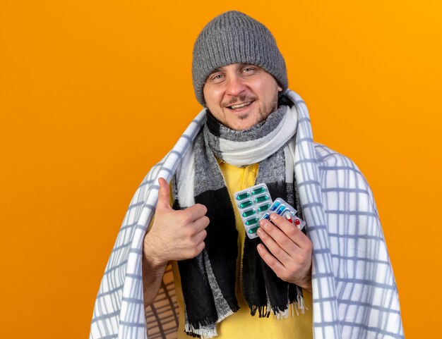 Feliz joven rubio enfermo eslavo con gorro de invierno y bufanda envuelto en cuadros pulgares hacia arriba y sostiene paquetes de píldoras médicas aisladas en la pared naranja con espacio de copia