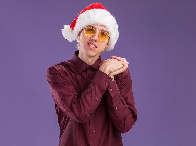 Feliz joven rubio con gorro de Papá Noel y gafas mirando a la cámara manteniendo las manos juntas aisladas sobre fondo púrpura con espacio de copia