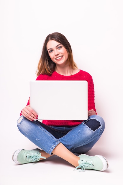 Foto gratuita feliz joven sentada en el suelo con las piernas cruzadas y usando la computadora portátil en la pared gris