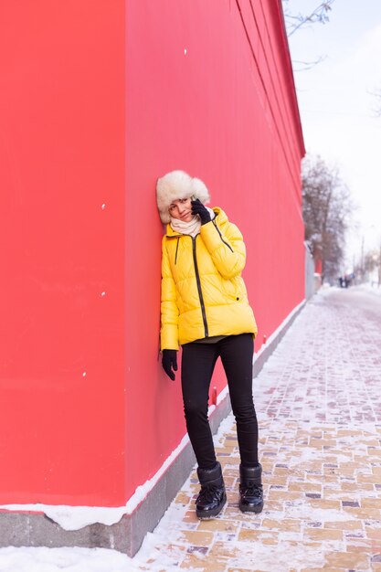 Feliz joven sobre un fondo de una pared roja en ropa de abrigo en un día soleado de invierno sonriendo y hablando por teléfono en una acera nevada de la ciudad