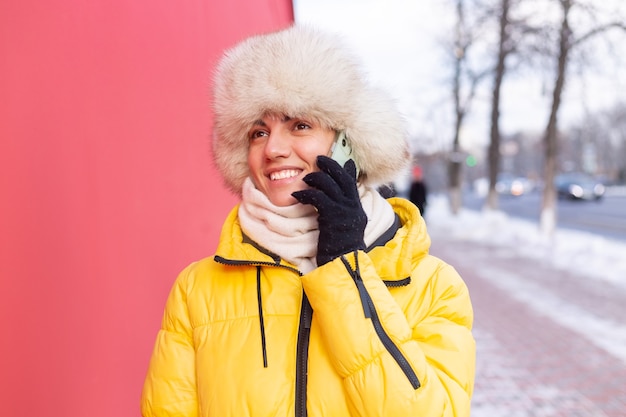 Foto gratuita feliz joven sobre un fondo de una pared roja en ropa de abrigo en un día soleado de invierno sonriendo y hablando por teléfono en una acera nevada de la ciudad