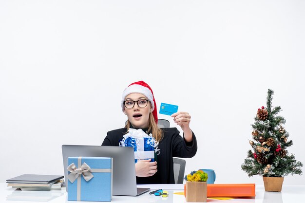 Feliz joven con sombrero de santa claus y gafas sentado en una mesa con regalo de Navidad y mirando la tarjeta bancaria sobre fondo blanco.
