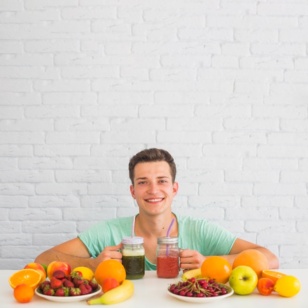 Feliz joven sosteniendo batidos con coloridas frutas orgánicas frescas en el escritorio