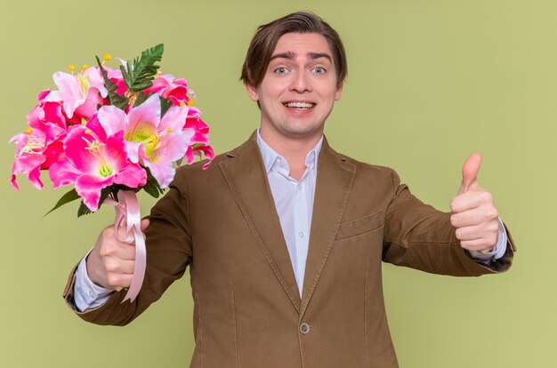 Feliz joven sosteniendo un ramo de flores sonriendo alegremente mostrando los pulgares hacia arriba para felicitar el día internacional de la mujer de pie sobre la pared verde