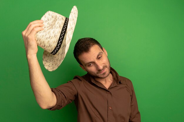 Feliz joven sosteniendo un sombrero de playa por encima de la cabeza mirando hacia abajo aislado en la pared verde