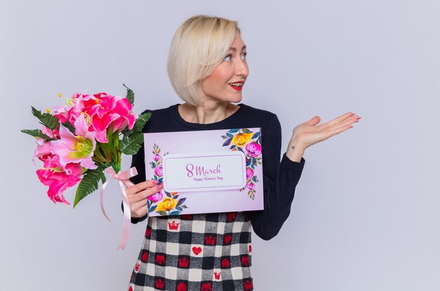 Feliz joven sosteniendo una tarjeta de felicitación y un ramo de flores mirando a un lado presentando algo con el brazo sonriendo celebrando la marcha del día internacional de la mujer