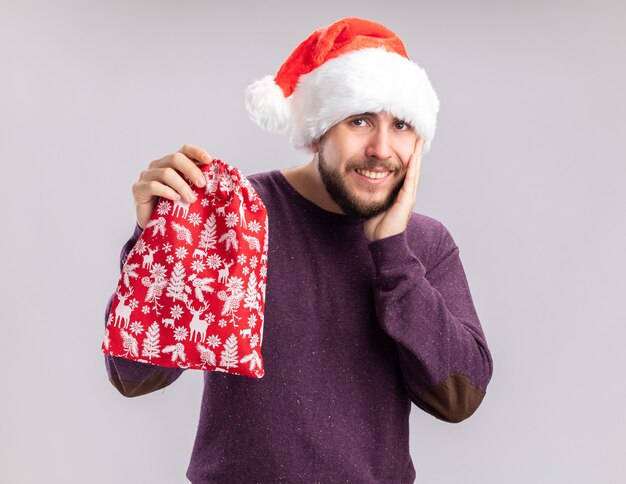 Feliz joven en suéter morado y gorro de Papá Noel con bolsa roja con regalos mirando a la cámara sonriendo alegremente de pie sobre fondo blanco.