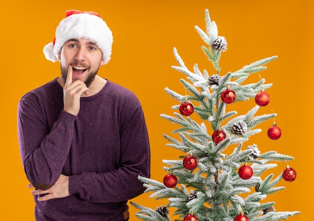 Feliz joven en suéter morado y gorro de Papá Noel mirando a la cámara con una sonrisa en la cara junto al árbol de Navidad sobre fondo naranja
