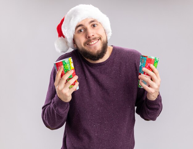 Feliz joven en suéter morado y sombrero de santa sosteniendo coloridos vasos de papel mirando a la cámara sonriendo alegremente de pie sobre fondo blanco.