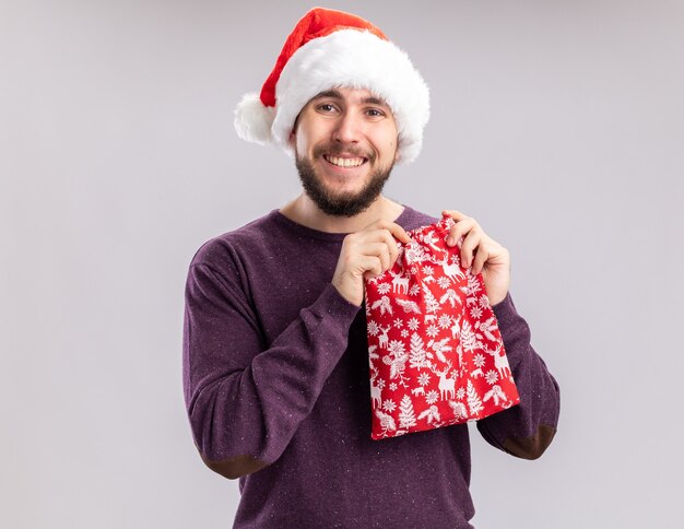 Feliz joven en suéter púrpura y gorro de Papá Noel con bolsa roja con regalos mirando a la cámara sonriendo ampliamente de pie sobre fondo blanco.