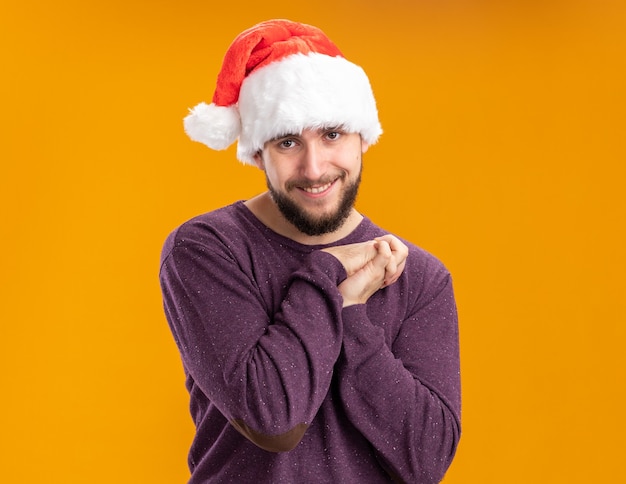 Feliz joven en suéter púrpura y gorro de Papá Noel cogidos de la mano juntos mirando a la cámara con una sonrisa en la cara de pie sobre fondo naranja