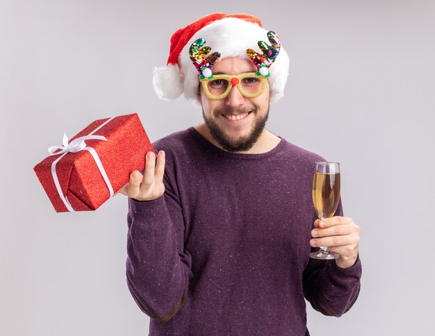 Feliz joven en suéter púrpura y gorro de Papá Noel con gafas divertidas sosteniendo una copa de champán y presente