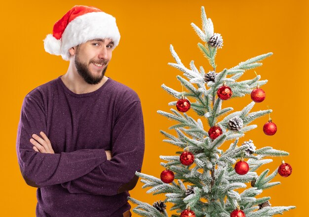 Feliz joven en suéter púrpura y gorro de Papá Noel mirando a la cámara sonriendo astutamente junto al árbol de Navidad sobre fondo naranja