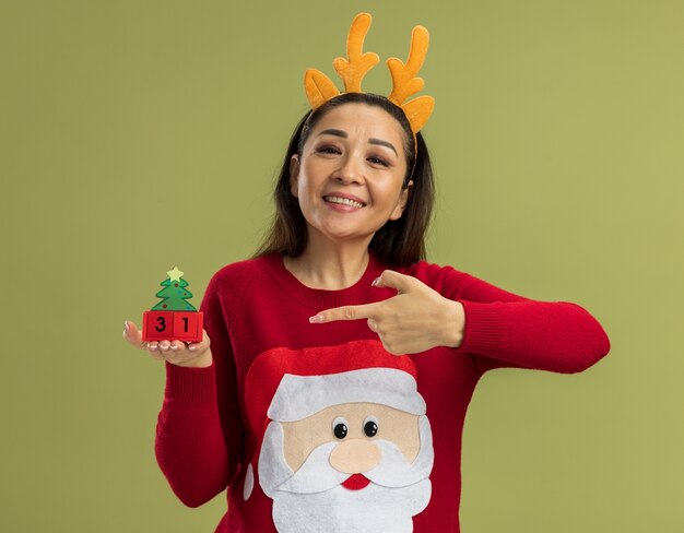 Feliz joven en suéter rojo de Navidad con borde divertido con cuernos de venado mostrando cubos de juguete con fecha de año nuevo sonriendo alegremente apuntando con el dedo índice a cubos de pie sobre fondo verde