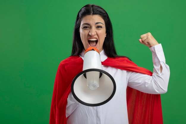 Feliz joven superhéroe caucásica chica gritando en altavoz y levantando un puño aislado en la pared verde con espacio de copia