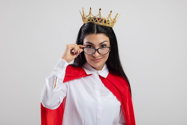 Feliz joven superhéroe caucásica con gafas y corona agarrando gafas mirando a cámara aislada sobre fondo blanco con espacio de copia