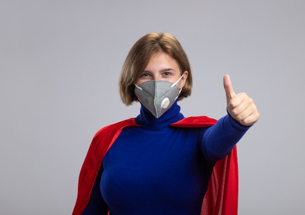 Feliz joven superhéroe rubia en capa roja con máscara protectora mirando a la cámara mostrando el pulgar hacia arriba aislado sobre fondo blanco con espacio de copia