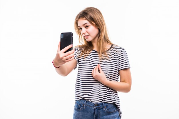 Feliz joven tomando fotos de sí misma en un teléfono inteligente, sobre una pared blanca