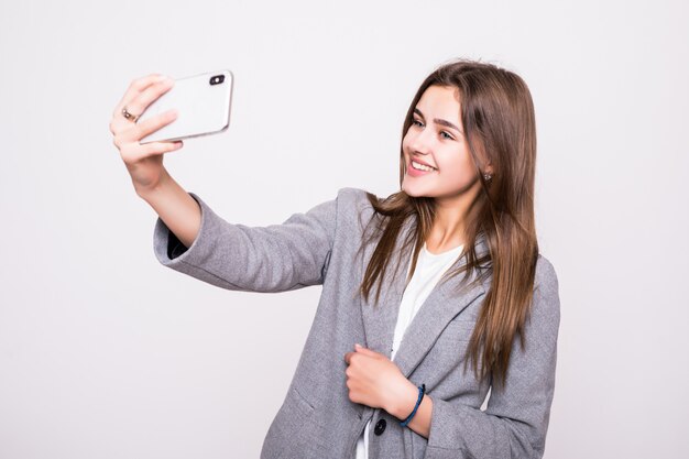 Feliz joven tomando fotos de sí misma a través del teléfono celular, sobre fondo blanco.