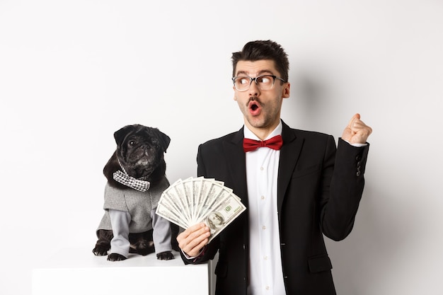 Feliz joven en traje ganar dinero con su perro. Guy regocijándose, sosteniendo dólares y mirando a la izquierda, pug negro en traje mirando a la cámara, fondo blanco.