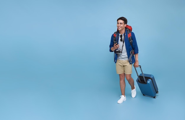 Foto gratuita feliz joven turista asiático sosteniendo un teléfono inteligente con equipaje que va a viajar de vacaciones aislado en el fondo del espacio de copia azul