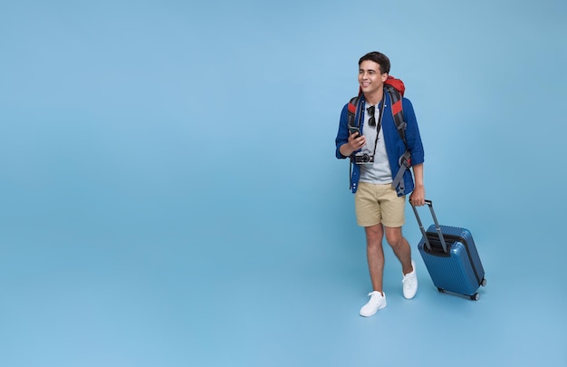 Feliz joven turista asiático sosteniendo un teléfono inteligente con equipaje que va a viajar de vacaciones aislado en el fondo del espacio de copia azul