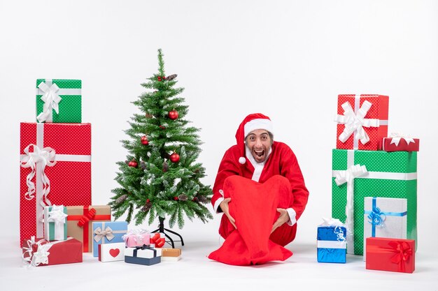 Feliz joven vestido como Papá Noel con regalos y árbol de Navidad decorado sentado en el suelo sobre fondo blanco.