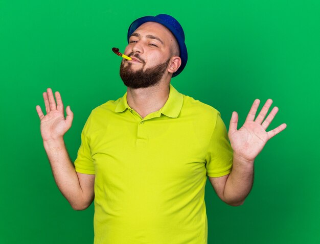 Feliz joven vestido con gorro de fiesta azul soplando silbato de fiesta extendiendo las manos