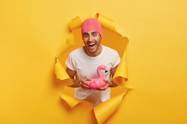 Feliz joven viste un gorro de baño de goma rosa, viste una camiseta blanca, sostiene un anillo de natación en forma de flamenco