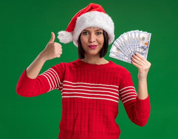 Feliz joven vistiendo gorro de Papá Noel con dinero mostrando el pulgar hacia arriba aislado en la pared verde