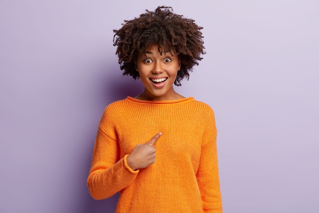 Feliz jovencita de piel oscura se señala a sí misma, se regocija de ser elegida, viste un jersey casual naranja, sonríe, aislada sobre una pared púrpura. Una mujer optimista y sonriente dice que te refieres a mí.