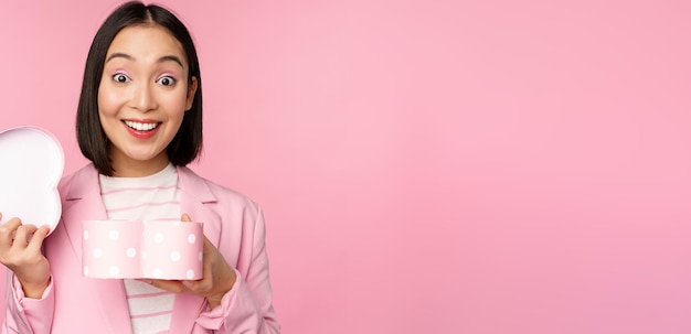 Feliz linda chica coreana en traje abre una caja en forma de corazón con un regalo romántico en las vacaciones del día blanco de pie en traje sobre fondo rosa