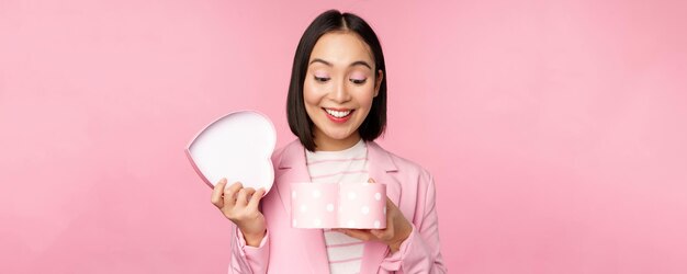 Feliz linda chica coreana en traje abre una caja en forma de corazón con un regalo romántico en las vacaciones del día blanco de pie en traje sobre fondo rosa